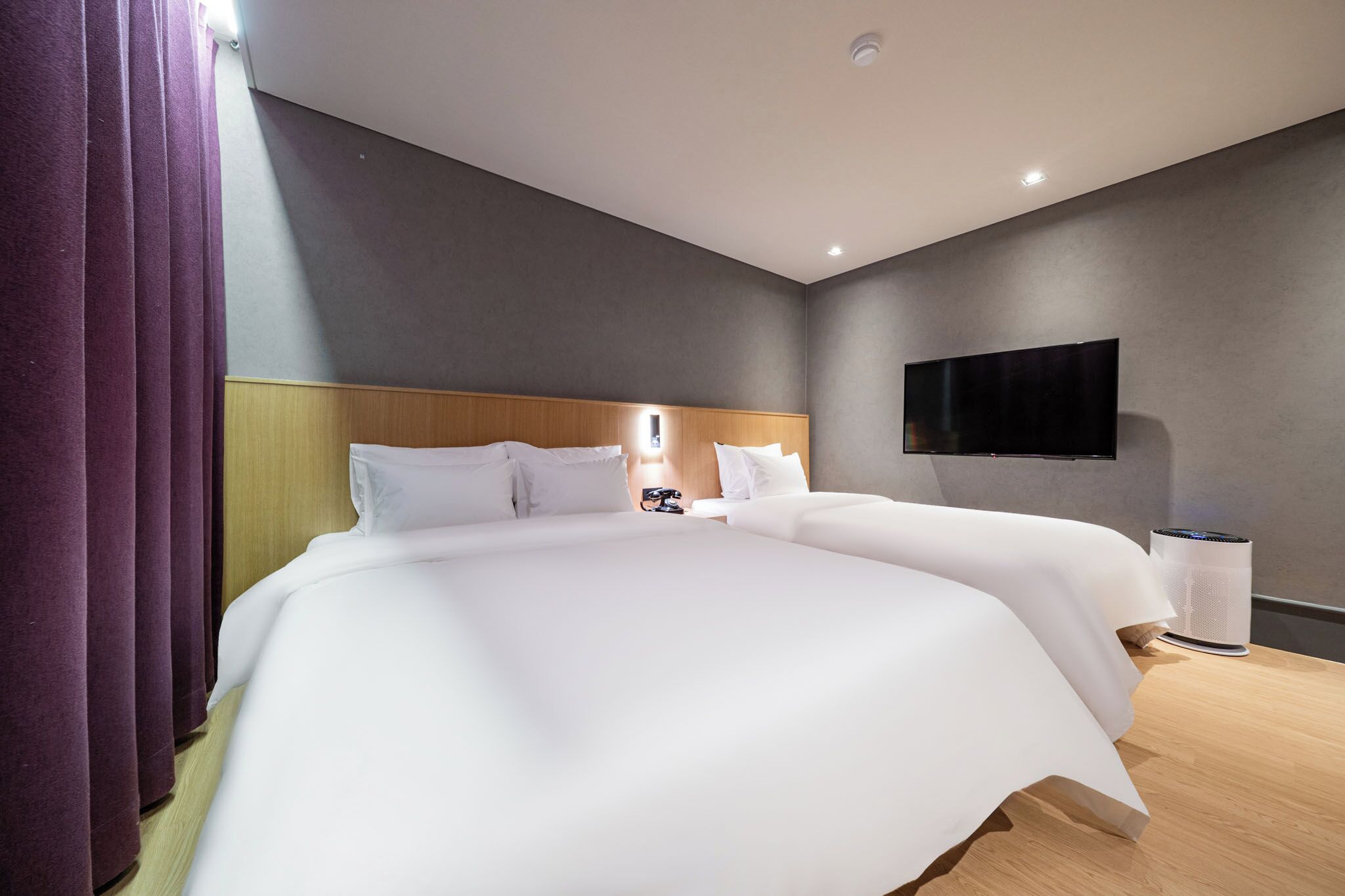 A modern hotel room with a large bed, a flat-screen TV mounted on the wall, and a bedside table with a lamp.