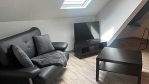 Ein modernes Wohnzimmer mit einer schwarzen Ledersofa, einem Flachbildfernseher auf einem Standfuß und einem kleinen Couchtisch. Ein Oberlicht lässt natürliches Licht herein.