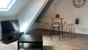 Ein Loft-Wohnzimmer mit schrägverdeckter Decke, einem schwarzen Sofa, einem kleinen Tisch und drei Hocker. Eine große Wanduhr und ein Flachbildfernseher sind zu sehen.