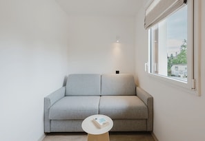A corner room with a grey sofa, a small white table, and a window with a view of buildings and trees. - 101 - Appart'Hôtel Neostelia-Annecy (Annecy)