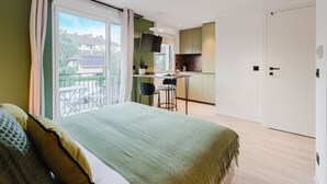 Ein modernes Schlafzimmer mit einem großen Bett, einem Balkon mit Blick auf Häuser, einer kleinen Küchenzeile und einer Schiebetür.