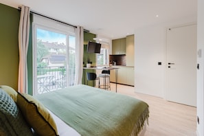 A modern bedroom with a large bed, a balcony with a view of houses, a small kitchen area, and a sliding door. - 103 - Appart'Hôtel Neostelia-Annecy (Annecy)