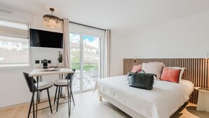 A modern bedroom with a large bed, a desk with two chairs, a TV mounted on the wall, and a balcony with a view of the neighborhood.