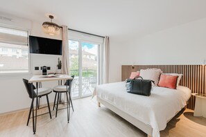 Un dormitorio moderno con una cama grande, un escritorio con dos sillas, un televisor montado en la pared y un balcón con vistas al vecindario.