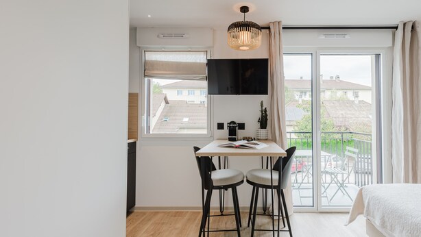 A modern bedroom with a large bed, a desk with two chairs, a TV, and a balcony with outdoor furniture.