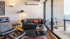 Un salón moderno con un sofá de cuero negro, una mesa de centro redonda con jarrones decorativos, una lámpara de pie negra y un ventanal con un equipo de aire acondicionado.