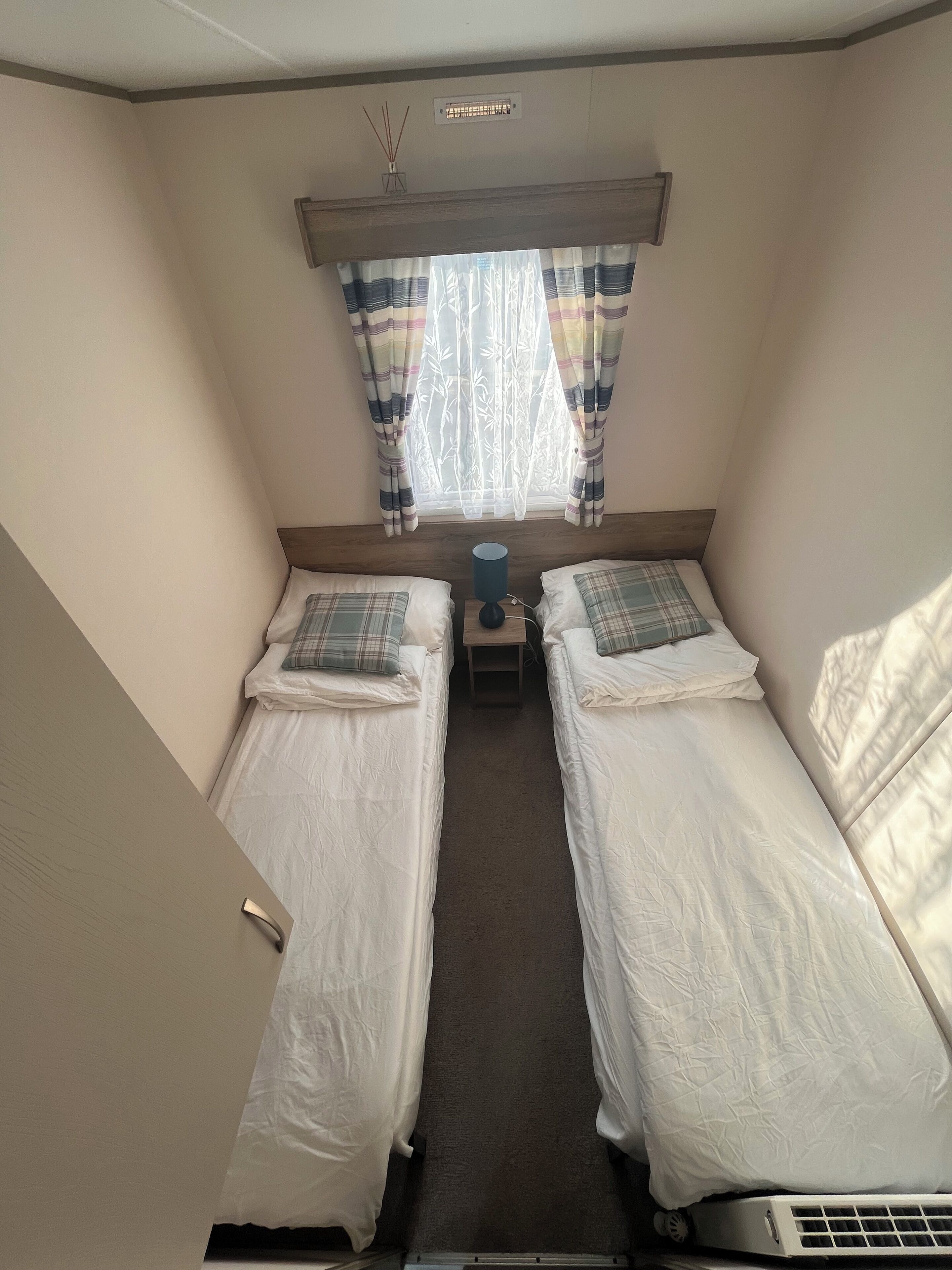 A small, compact room with two single beds, a bedside table with a lamp, a window with curtains, and a radiator.