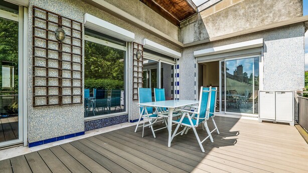 Un patio al aire libre con un suelo de madera, una mesa con sillas azules y una puerta de cristal que da a un jardín.