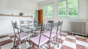 Comedor con mesa de vidrio y sillas metálicas, ventanas amplias con vista a vegetación, un radiador y un estante de madera con plantas.