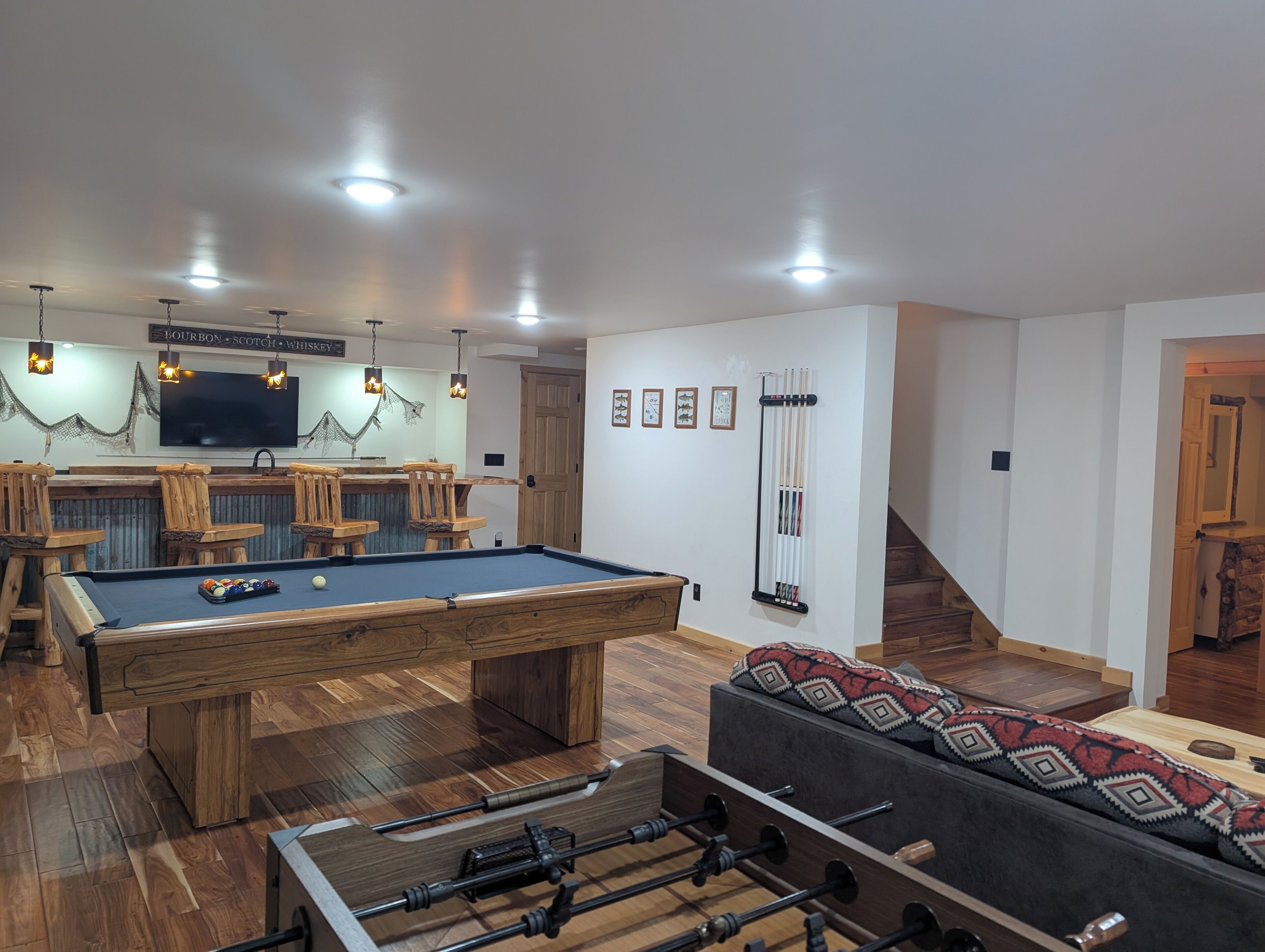 A pool table, bar area, and a sofa in a room with wooden flooring and white walls.