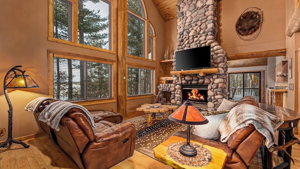 A cozy living room with a stone fireplace, leather furniture, and large windows offering a view of the forest.