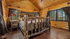 A log cabin bedroom with a rustic bed frame, a ceiling fan, and large windows offering a forest view.