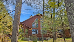 A log cabin with a large window and a stone chimney, surrounded by trees and a grassy area with steps leading to the entrance.