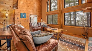 A cozy cabin interior with a leather sofa, a wooden dining table, and large windows offering a view of the forest.