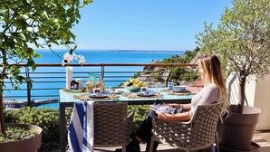 Iemand zit aan een tafel met uitzicht op zee, een bloempot en een houten terras.