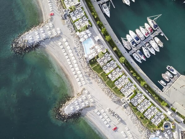 An aerial view of a marina with boats docked, a sandy beach with lounge chairs, and a pool area.