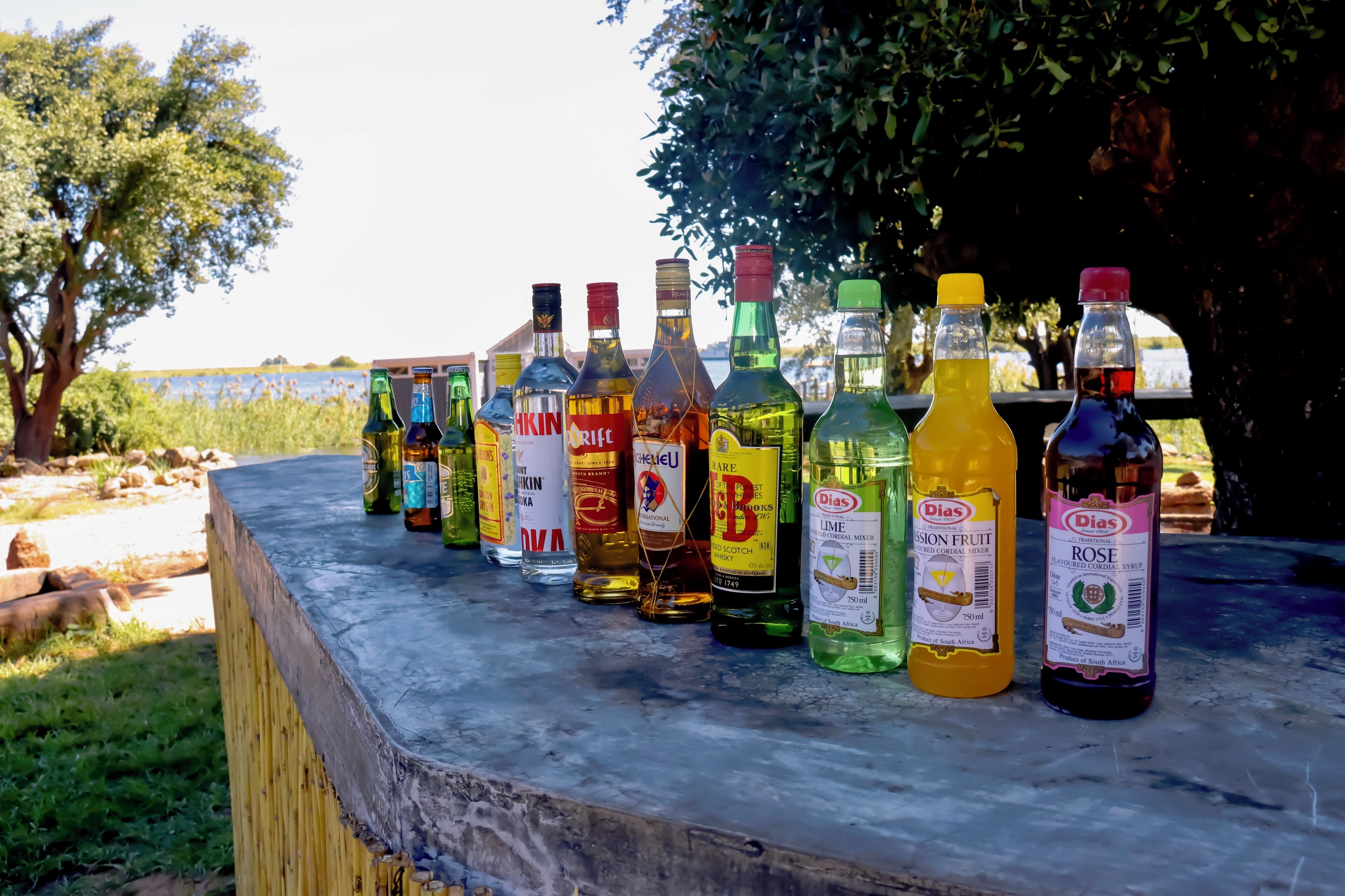 Varias botellas de licores alineadas sobre un repisa de madera junto al agua.