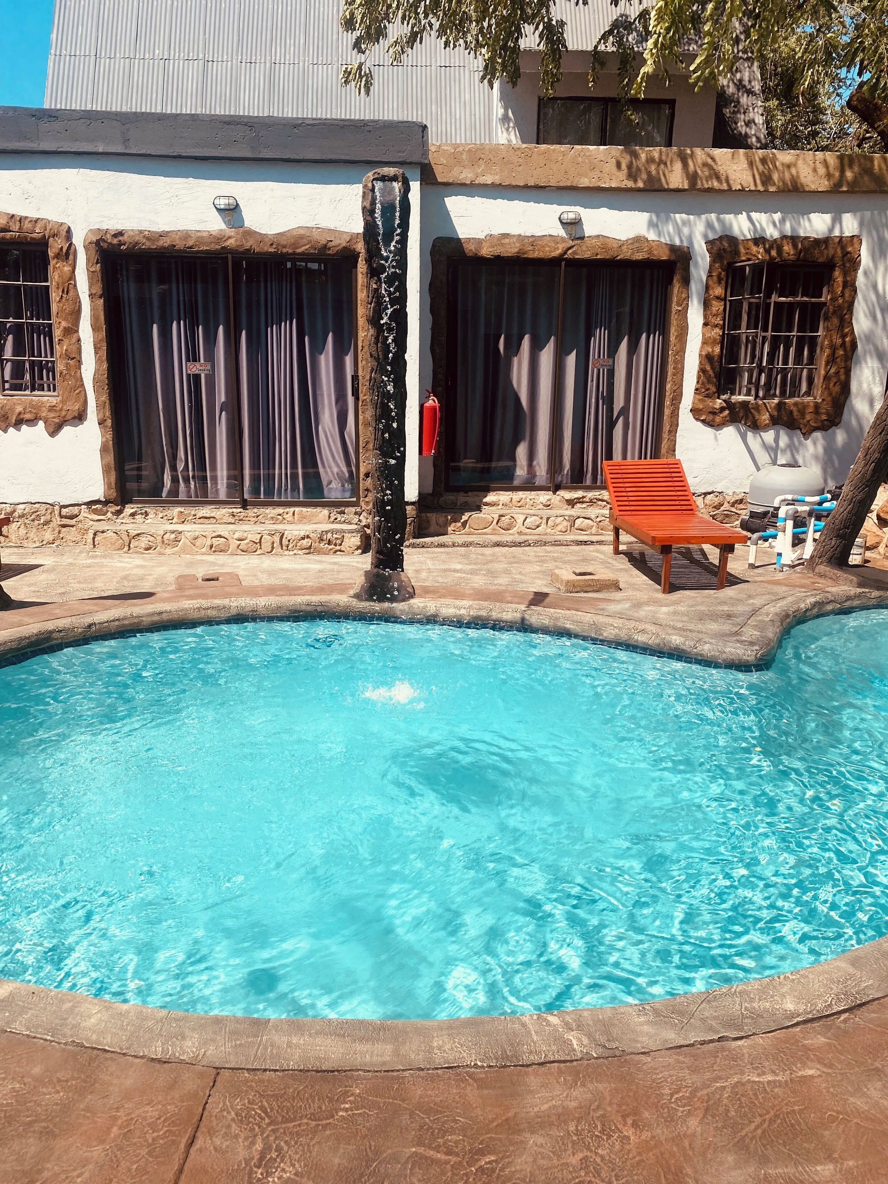 Una piscina con una silla de madera, un extintor y un edificio de piedra rústica y ventanas grandes.