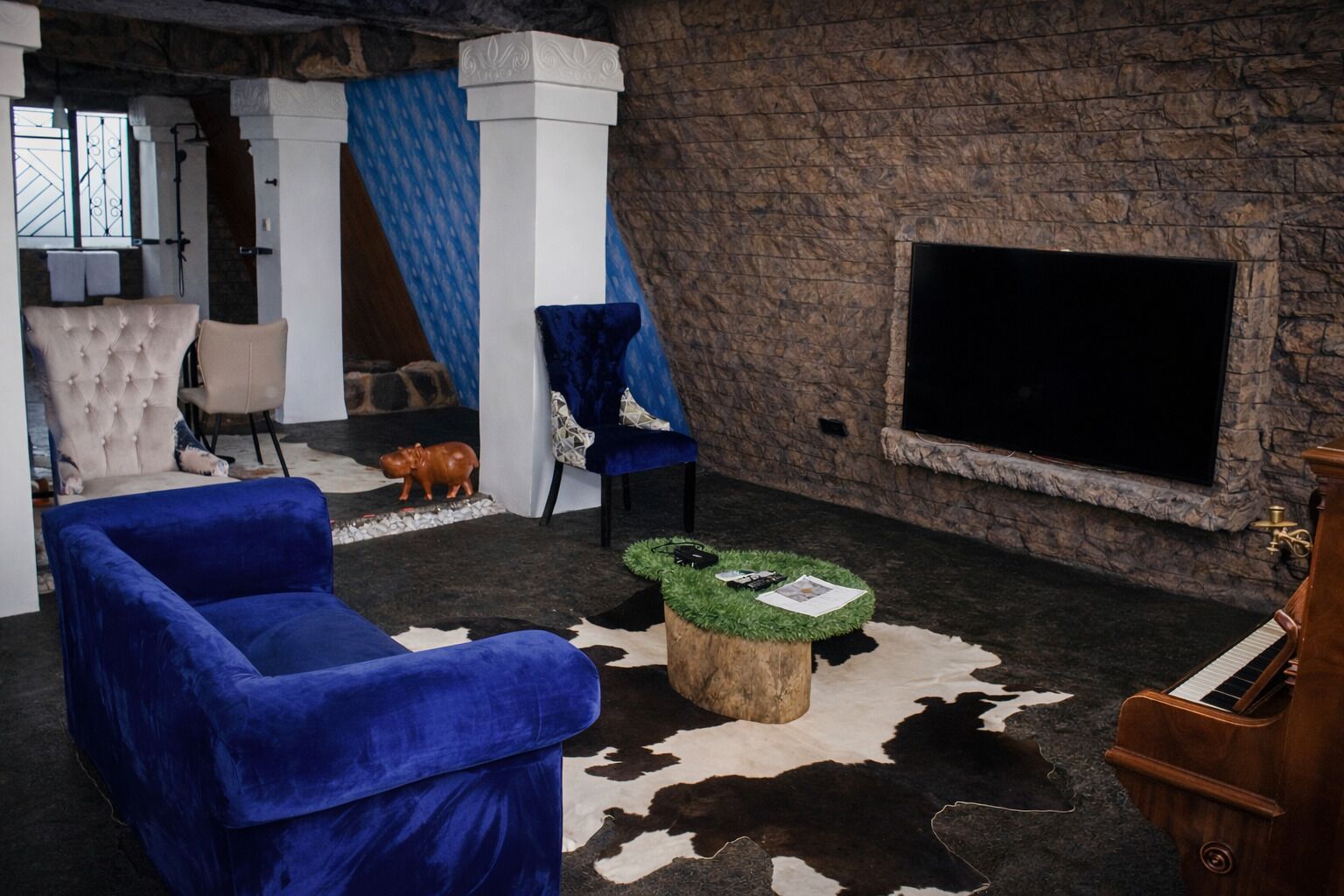 A room with a large TV, a blue armchair, a tufted armchair, a small wooden table with a cowhide rug, and a piano.
