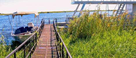 Un muelle metálico que conduce a un bote, rodeado de hierba alta y un cielo azul despejado.