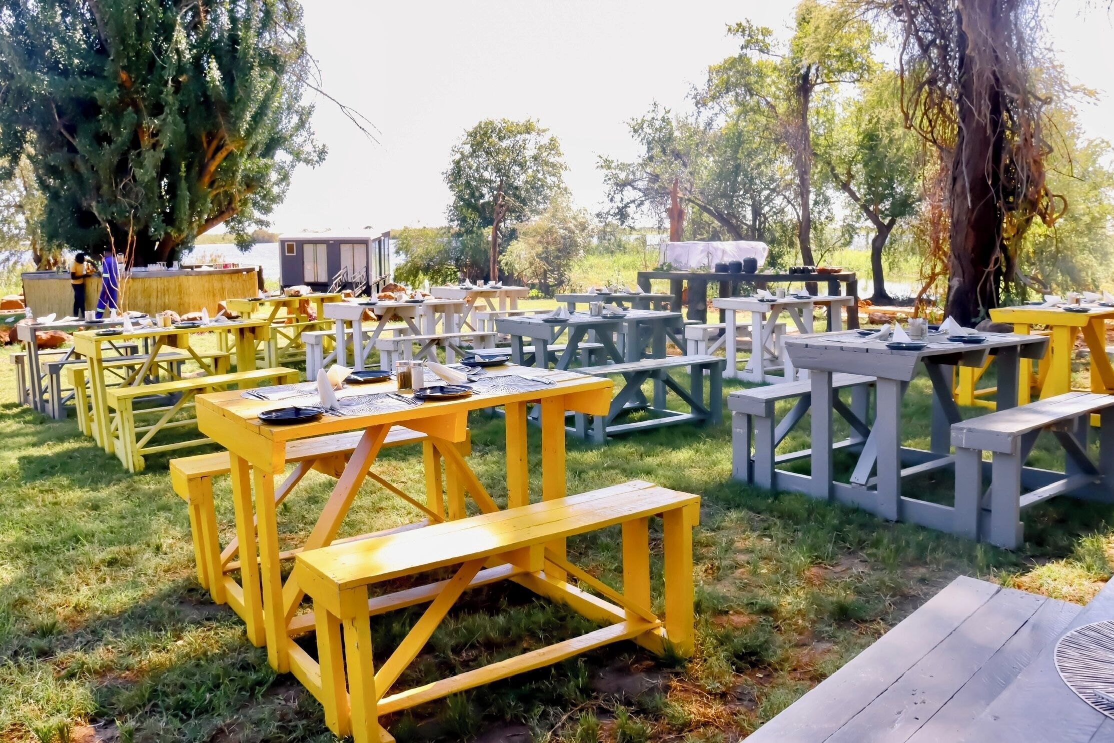 Un espace de restauration en plein air doté de plusieurs tables et bancs en bois, entouré d’arbres et bordé par un plan d’eau.