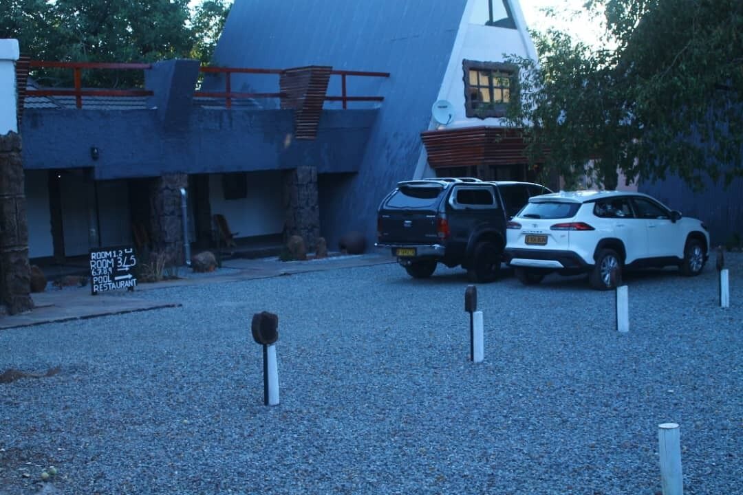 Un edificio con un diseño arquitectónico singular, que incluye un techo inclinado y un ventanal grande. Hay dos coches aparcados delante, y un cartel que indica la disponibilidad de habitaciones y piscina.