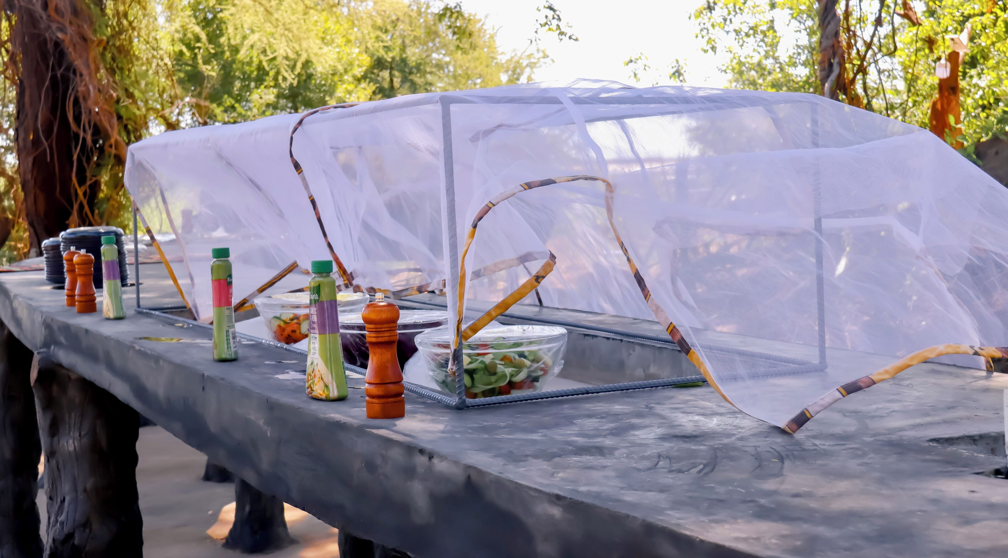 Una mesa con una cubierta transparente, varias botellas de condimentos y un recipiente con ensalada.