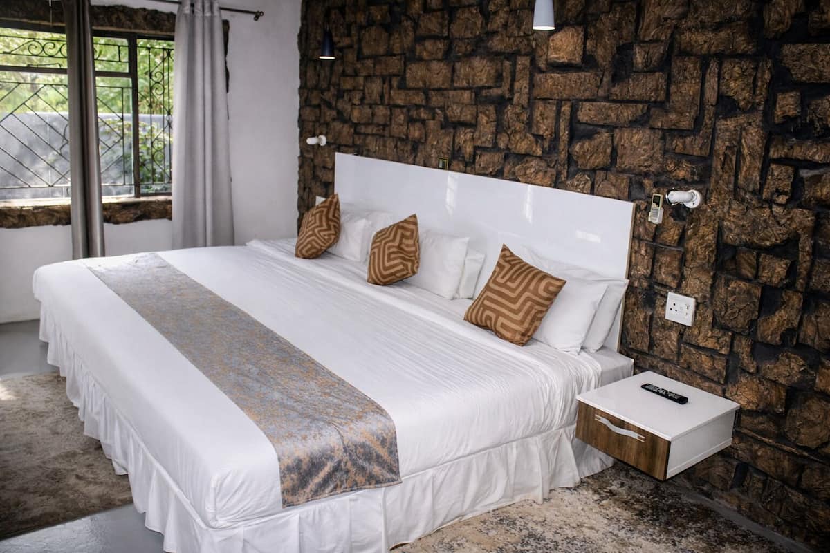 A bedroom with a stone wall, a white bed with patterned pillows, a bedside table with a remote control, and a window with curtains.