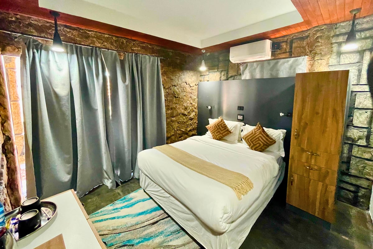 A bedroom with a bed, curtains, a headboard, a wooden wardrobe, and a stone wall.