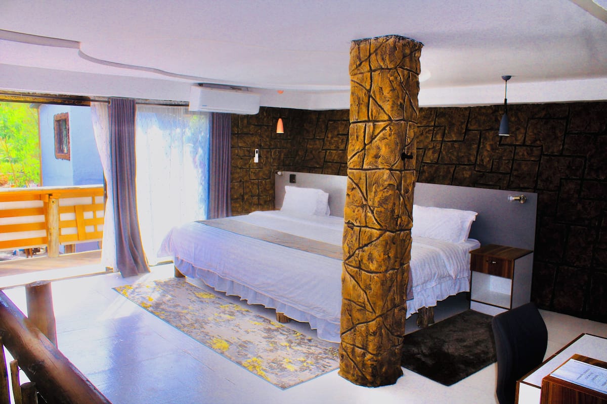 A bedroom with a large bed, a stone pillar, a desk with a chair, a nightstand, and a view of the outside.