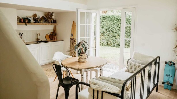 A kitchen with white cabinets, a dining table with chairs, and a view of a garden through a glass door. - Maison de Vacance en Lisière de Forêt (Labenne)