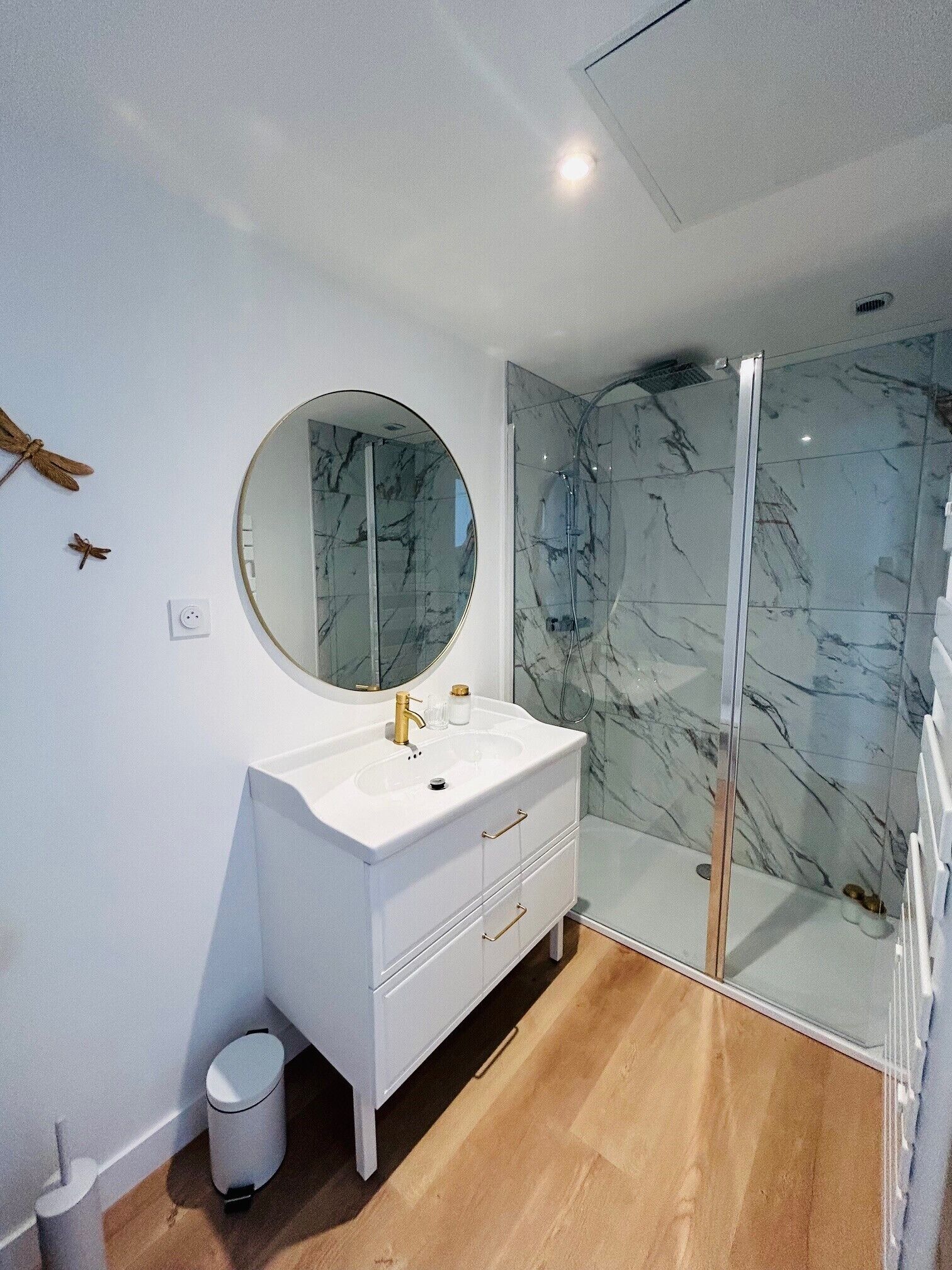A modern bathroom with a white vanity, a round mirror, a glass shower enclosure, and marble tiles.