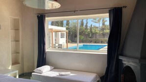A room with a bed, a window with a view of a pool, a fireplace, and a ceiling with exposed beams. - Villa Mogador Essaoura (Essaouira)