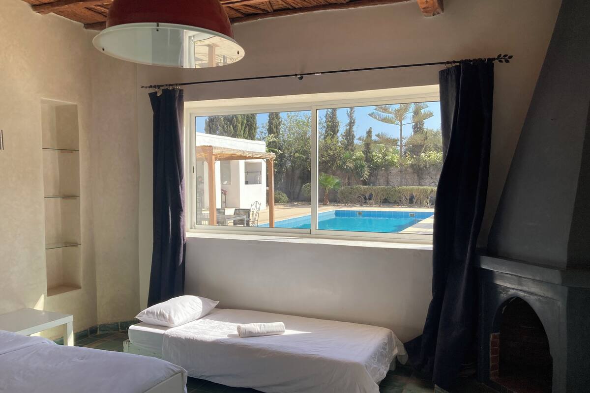 A room with a bed, a window with a view of a pool, a fireplace, and a ceiling with exposed beams.