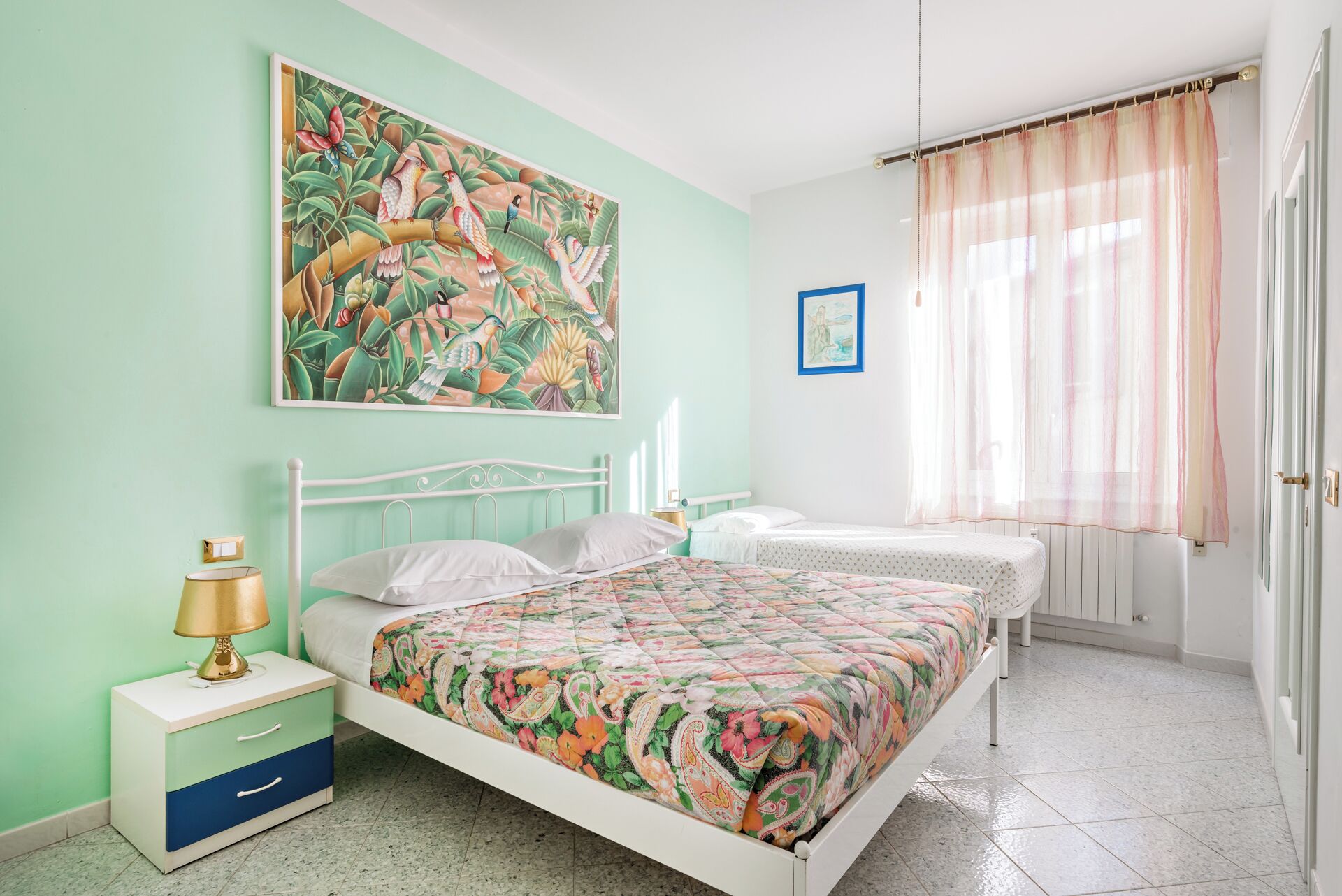 A bedroom with a bed, bedside tables, a nightstand, a painting, and a window with curtains.