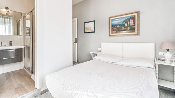 Une chambre avec un grand lit, une table de chevet avec une lampe, un tableau au mur et une salle de bain avec une douche et un miroir.