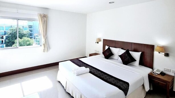 A hotel room with a large bed, bedside tables, a window with curtains, and a view of buildings outside. - Siamese Serenity Stays (Pattaya)