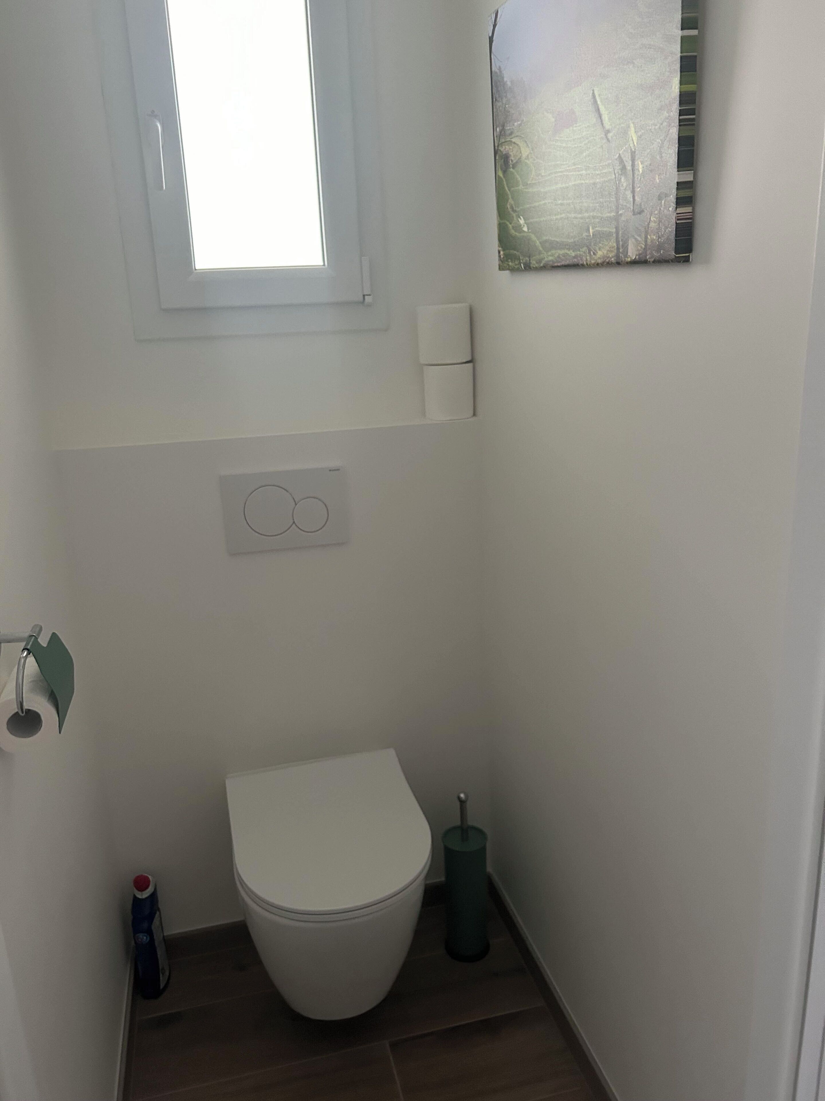 Een kleine, witte badkamer met toilet, raam, wandlamp en prullenbak.