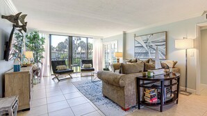 A spacious living room with a large window, a sofa, a coffee table, and a bookshelf. - Luxe Fort Pierce Beach Condo w/ Golf Views! (Fort Pierce)
