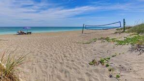 A sandy beach with a volleyball net and a person relaxing under a purple umbrella. - Luxe Fort Pierce Beach Condo w/ Golf Views! (Fort Pierce)