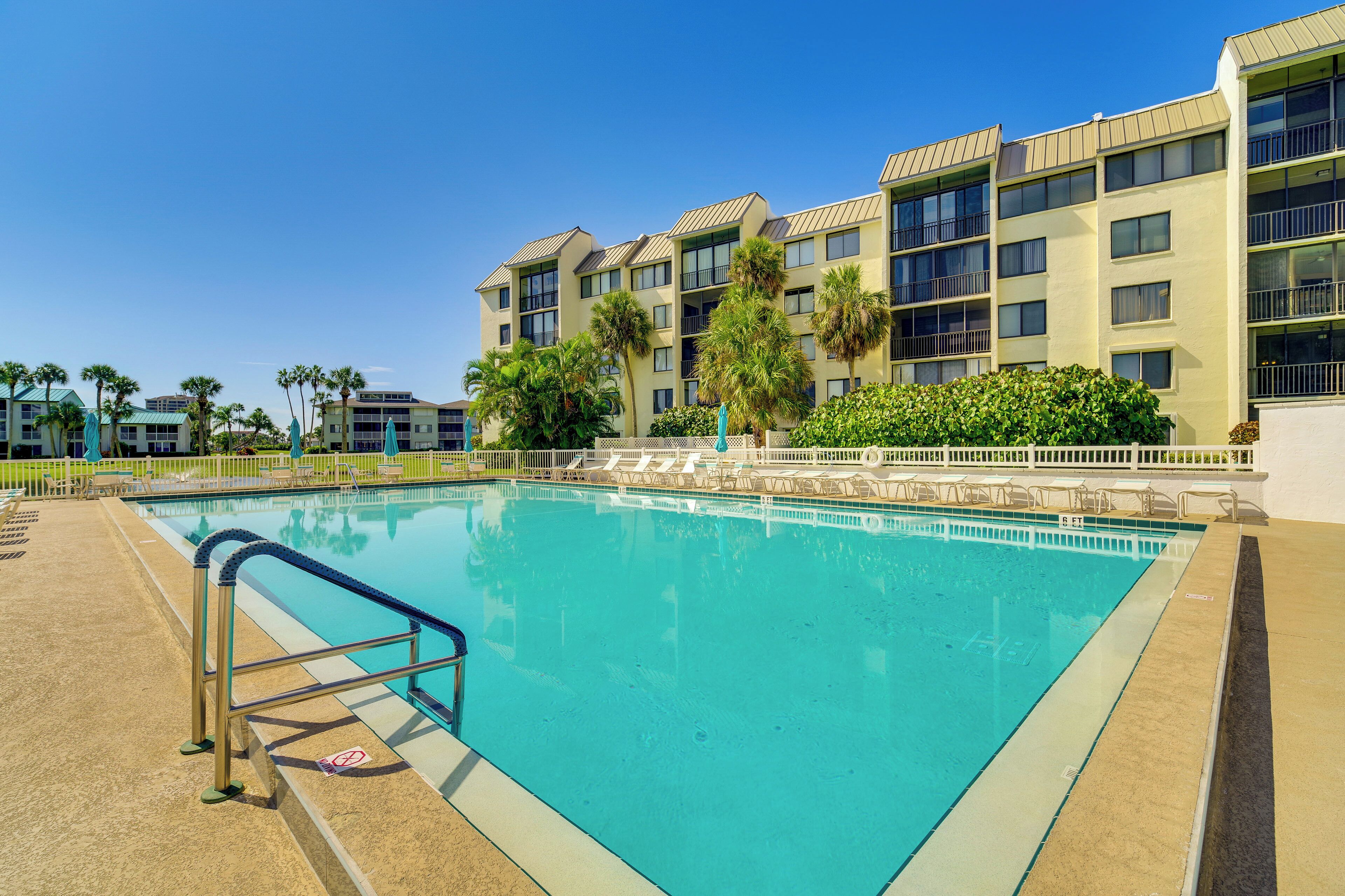 A swimming pool with a metal ladder, surrounded by a fence, and a building with multiple balconies in the background.