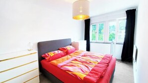 Un dormitorio con cama, mesita de noche, lámpara y ventana con cortinas.
