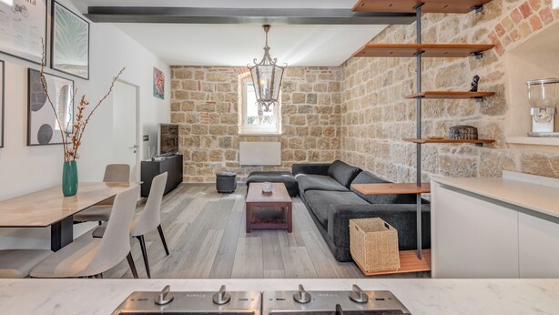 A modern living room with a stone wall, a sofa, a dining table, and a kitchen area with wooden shelves. - Funky Donkey (Split)
