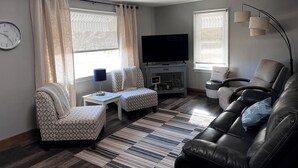 A living room with a sofa, armchair, ottoman, TV, and a small table with a lamp.
