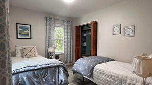 A bedroom with two beds, a wooden floor, a window with curtains, and a small table with books.