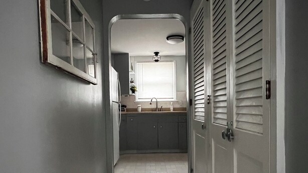A narrow hallway with a kitchen at the end, featuring a sink, cabinets, and a window with blinds.