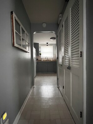 A narrow hallway with a kitchen at the end, featuring a sink, cabinets, and a window with blinds.
