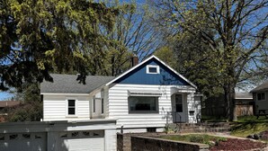 A two-story house with a blue roof and white siding, a front garage, and a stone retaining wall with a small garden.