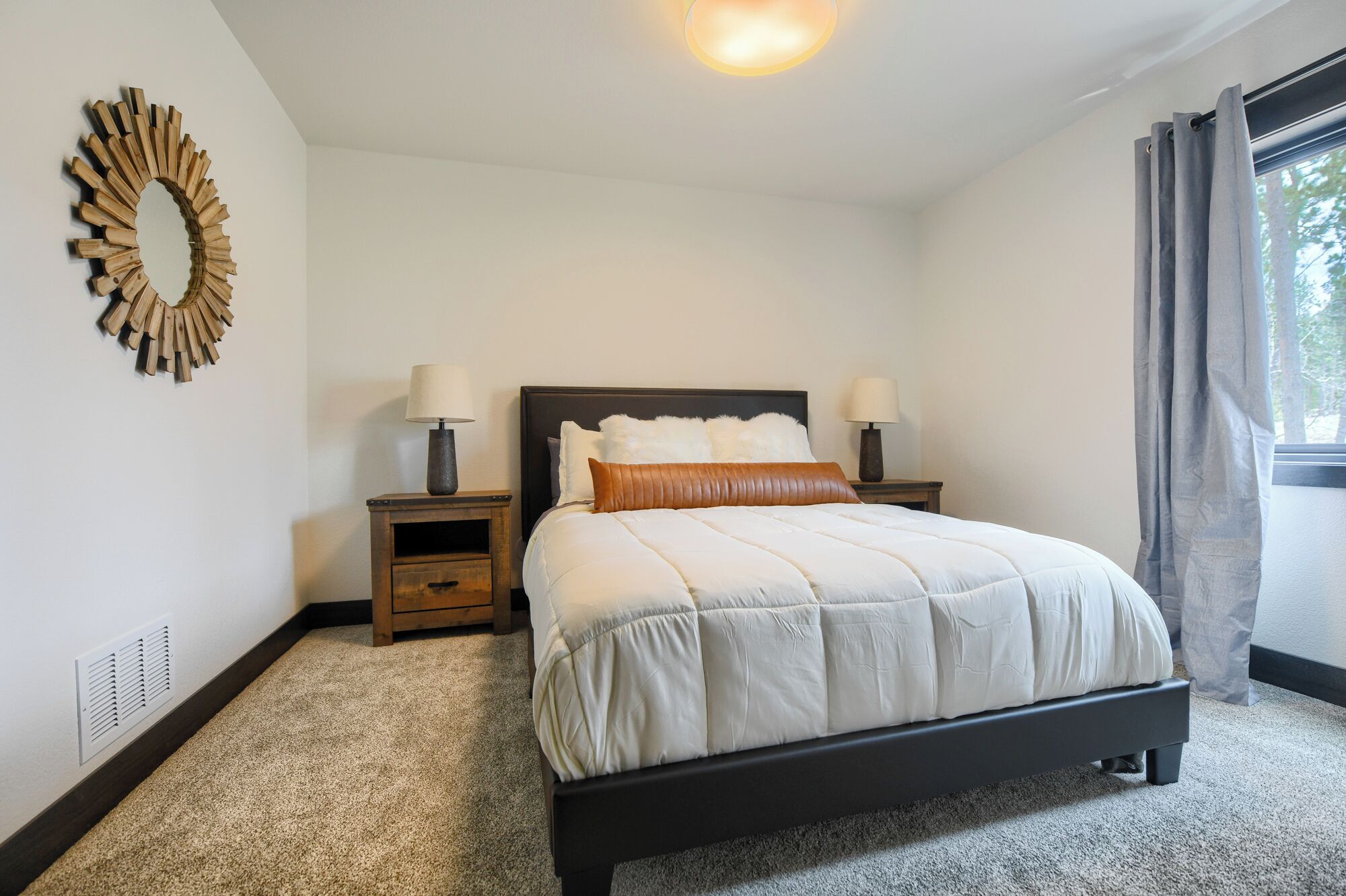 Un dormitorio con cama, dos mesitas de noche con lámparas, un espejo en la pared y una ventana con cortinas.