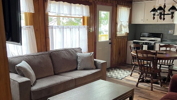 Ein gemütliches Wohnzimmer mit einer Couch, einem Couchtisch und einem roten Sofa. Im Hintergrund ist eine Küche mit einem Herd und Schränken zu sehen.
